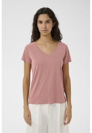 Vrouw met golvend donker haar, gekleed in een stofroze T-shirt met V-hals en korte mouwen en losse witte broek, staand tegen een effen achtergrond.