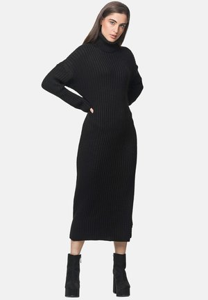 Robe noire à col roulé côtelé avec des manches longues, longueur mi-mollet, silhouette ajustée et fentes latérales ; associée à des bottines noires.