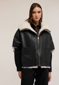 Kobieta w czarnej skórzanej kurtce z białym kołnierzem i lamówką ze sztucznego futra, zapięta, na neutralnym tle.