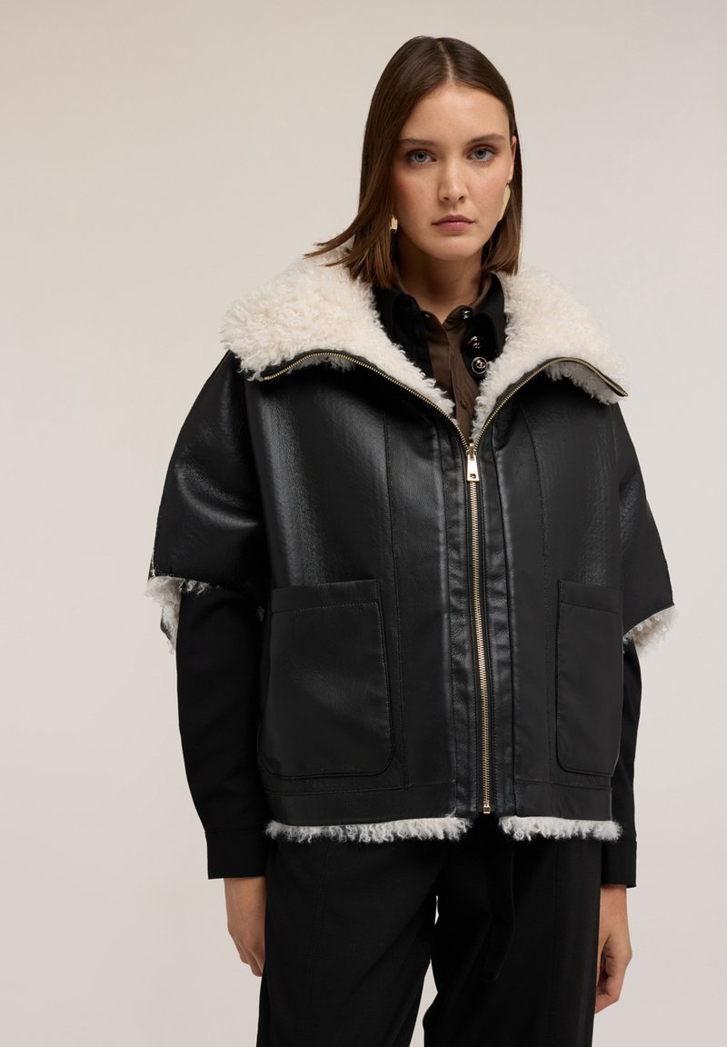 Kobieta w czarnej skórzanej kurtce z białym kołnierzem i lamówką ze sztucznego futra, zapięta, na neutralnym tle.