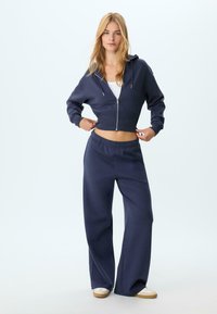 Felpa croppata blu navy con zip frontale e cordino, abbinata a pantaloni felpati a gamba larga coordinati. Tessuto morbido, design casual.