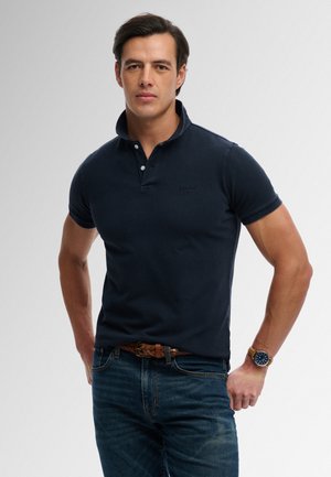 Uomo che indossa una polo blu scuro, jeans blu con cintura marrone intrecciata e un orologio da polso, in piedi con le mani sui fianchi.