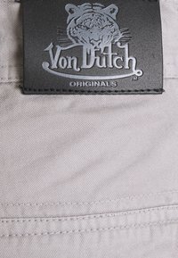 Melna ādas birka ar pelēku izspiestu tīģera grafiku, uz kuras ir teksts "Von Dutch" un "ORIGINALS". Pievienota gaismas pelēkam audumam ar smalku struktūru.