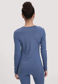 Vrouw met krullend haar in een knot, gekleed in een aansluitend blauw long-sleeve shirt en bijpassende blauwe legging, staand met haar rug naar de camera.