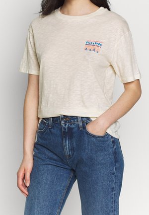 Kobieta nosząca jasnobeżowy t-shirt z krótkim rękawem z małą kolorową grafiką oraz niebieskie jeansy z wysokim stanem, jedna ręka w kieszeni.