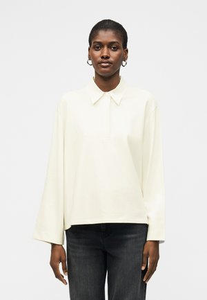 Γυναίκα με κοντά μαλλιά που φοράει μεγάλα σκουλαρίκια κρίκους, ένα άνετο λευκό πουκάμισο με μακριά μανίκια και γιακά και μαύρο τζιν μπροστά σε απλό φόντο.