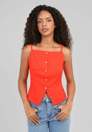 Femme aux cheveux bouclés et foncés portant un haut sans manches orange vif à boutons et un jean bleu clair, les mains dans les poches.