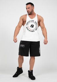 Witte tanktop met zwart cirkelvormig logo en tekst, gecombineerd met zwarte shorts met een logopatch en zijaccenten. Zwarte schoenen.