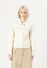 Veste en denim crème avec col, boutonnière à l'avant, deux poches poitrine et manches longues. Portée sur une jupe beige. Fond uni.