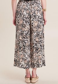 Palazzo-broeken in zwart en wit met een roze tropisch bladpatroon. Elastische tailleband en wijde pijpen. Het model draagt platform sandalen.