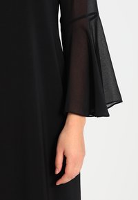 Robe noire avec des manches transparentes en forme de cloche. Le tissu est lisse, avec une texture fluide et une coupe ajustée.