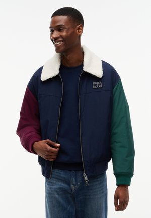 Homme souriant portant une veste color-block avec un col en shearling blanc, corps bleu marine, manche gauche bordeaux et manche droite verte.