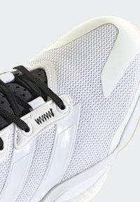 Zapatilla deportiva blanca con parte superior de malla, cordones negros y acentos sintéticos brillantes. Suela texturizada y detalle distintivo de diseño con tres rayas.