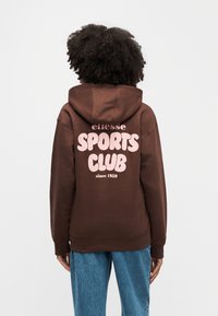Personne aux cheveux bouclés portant un sweat à capuche marron avec le texte rose « ellesse SPORTS CLUB depuis 1959 » dans le dos et un jean bleu, vue de dos.