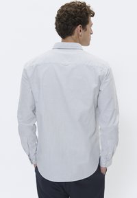 Mann mit lockigem braunem Haar, der ein hellblaues und weißes dünn gestreiftes Langarmhemd trägt, mit den Händen in den Taschen, von hinten auf einem einfarbigen Hintergrund.