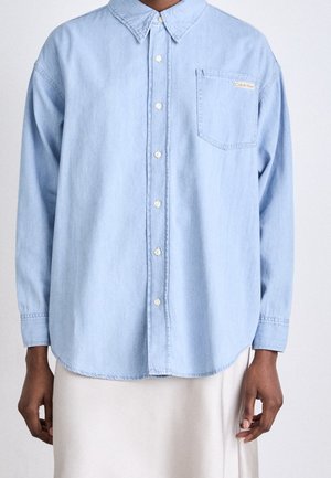 Lichtblauw overhemd met lange mouwen, knopen, kraag en borstzak, gedragen over een witte rok of broek.