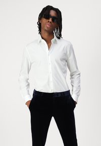 Biely dlhý rukáv košele s gombíkmi, slim fit, s špicatým golierom. Skombinované s tmavomodrými, jemne zamatovými nohavicami. Žiadne viditeľné vzory ani logá.