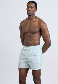 Μυώδης άνδρας που στέκεται με τα χέρια πίσω από την πλάτη, φοράει ανοιχτό μπλε μαγιό μπροστά σε απλό ανοιχτό φόντο.