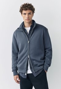 Donkergrijze zip-up sweater van zachte stof, met een hoge kraag en geribbelde manchetten. Hij is gelaagd over een blauw-wit gestreept shirt.