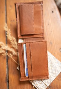 Portafoglio in pelle tan con tre slot per carte, texture liscia e cuciture visibili. Posizionato su una superficie decorata con accenti chiari.