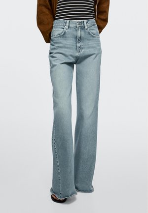 Lichtblauwe jeans met hoge taille en brede pijpen, gedragen met een zwart-wit gestreepte top en een bruine jas, gecombineerd met zwarte sandalen met open teen.