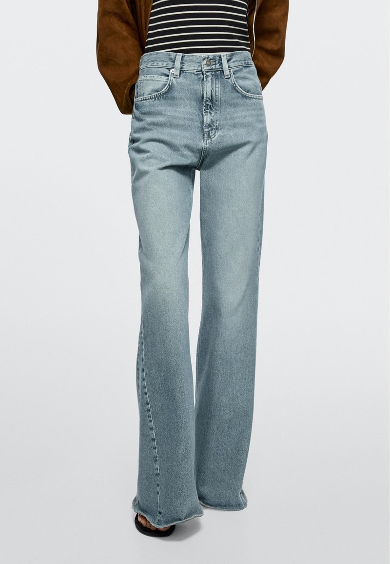 Lichtblauwe jeans met hoge taille en brede pijpen, gedragen met een zwart-wit gestreepte top en een bruine jas, gecombineerd met zwarte sandalen met open teen.