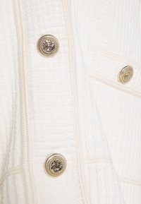 Kremowa tkanina o teksturze z złotymi, ozdobnymi guzikami z misternymi wzorami, widok z bliska na odzież.