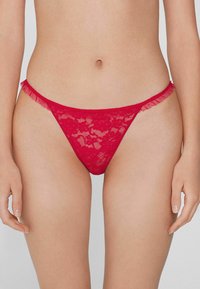 String rouge en dentelle avec un motif floral, présentant des accents froncés sur la taille et un design à couverture minimale. Texture lisse.