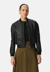 Giacca bomber nera in pelle con polsini e orlo a costine, zip anteriore e tasche laterali. Indossata sopra un top nero aderente e una gonna oliva.