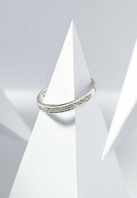 Silberner Ring mit einem glatten Band und einem strukturierten Abschnitt, der kleine klare Steine zeigt, der auf geometrischen weißen Papierstrukturen ruht.