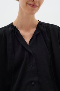 Černá lehká blůza s jemnou texturou, nařasenými rameny, výstřihem do V a zapínáním na malé černé knoflíky.