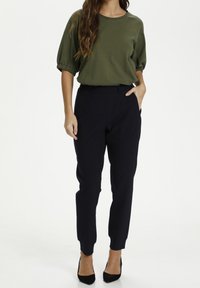 Vrouw in een olijfgroene blouse met korte mouwen en zwarte tapered broek met zwarte puntige hakken, staand met één hand in de zak.