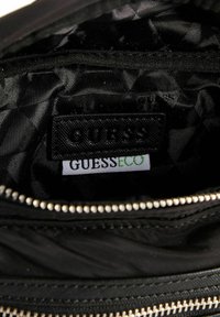 Intérieur matelassé noir avec une finition texturée. Comprend une étiquette "GUESS" et un tag "GUESS ECO". Fermeture éclair argentée avec des tirettes rondes.