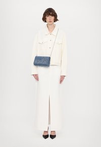Giacca in denim bianco con due tasche sul petto sopra un vestito maxi bianco con spacco frontale. Borsa in denim blu con lettere metalliche e tracolla a catena.