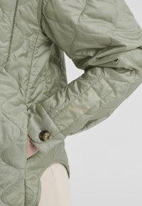 Quilted, hellgrüne Jacke mit sichtbaren Nähten, einem Knopfverschluss am Ärmel mit einem olivgrünen Knopf und einem strukturierten, glatten Stoff.