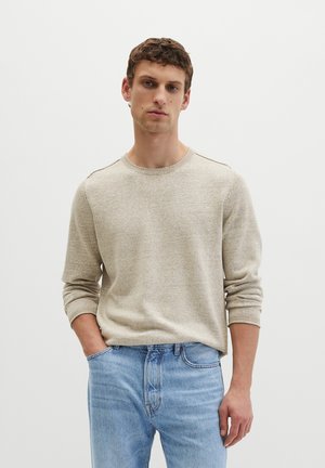 Giovane uomo che indossa un maglione beige girocollo e jeans azzurri chiaro, in piedi con una mano in tasca su uno sfondo bianco semplice.