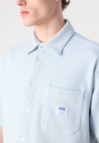 Camicia azzurro chiaro a maniche corte con una texture a coste, chiusura con bottoni, colletto e una tasca applicata con etichetta del marchio.