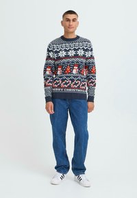Navy blue festive sweater with white, red, and patterned details including snowmen and candy canes. Worn with blue jeans and white sneakers.
