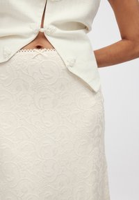 Blouse légère crème avec des boutons en spirale, associée à une jupe texturée crème présentant un motif floral complexe.