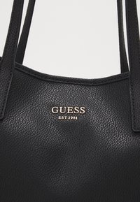 Sac fourre-tout en cuir noir texturé avec un détail en métal doré "GUESS EST 1981" à l'avant. Double poignées robustes pour le transport.