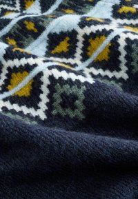 Adīta sega ar ģeometrisku rakstu tumši zilos, mentola zaļos, dzeltenos un baltos toņos, ar mīkstu tekstūru un bagātīgu krāsu kontrastu.