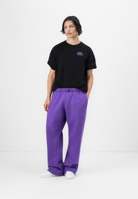 Svart bomull T-skjorte med rullede ermer og lilla brodert logo, kombinert med løstsittende lilla joggebukser og hvite sneakers.
