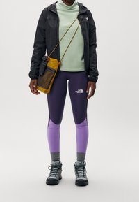 Chaqueta ligera negra, cuello alto verde menta, leggings morados y lilas, calcetines grises y botas de senderismo grises, con un bolso cruzado amarillo.