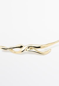 Fermaglio dorato con un design scultoreo e ondulato, caratterizzato da curve morbide e una finitura lucida. Lungo e sottile, senza ulteriori accenti.