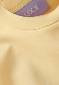 Lys gul bomull T-skjorte med ribbestrikket rund hals og en lilla etikett med teksten "JXXX" inne i kragen. Myk og glatt tekstur.