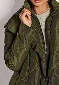 Olijfgroene pufferjack met hoge kraag, ritssluiting en een ruitpatroon. Gelaagd, glanzende stof met een warme uitstraling.