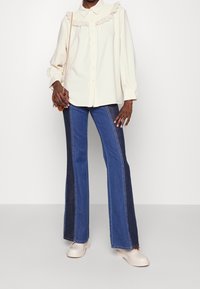 Person wearing a cream blouse with ruffled collar, blue two-tone flared jeans, cream shoes, and holding a small brown crossbody bag.