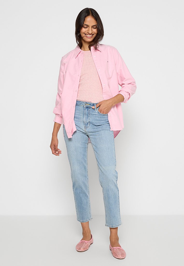 Jasnoróżowa koszula zapinana na guziki noszona na jasnoróżowym swetrze, w zestawie z jasnoniebieskimi dżinsami i różowymi butami typu slip-on. Prosty design, casualowy fason.
