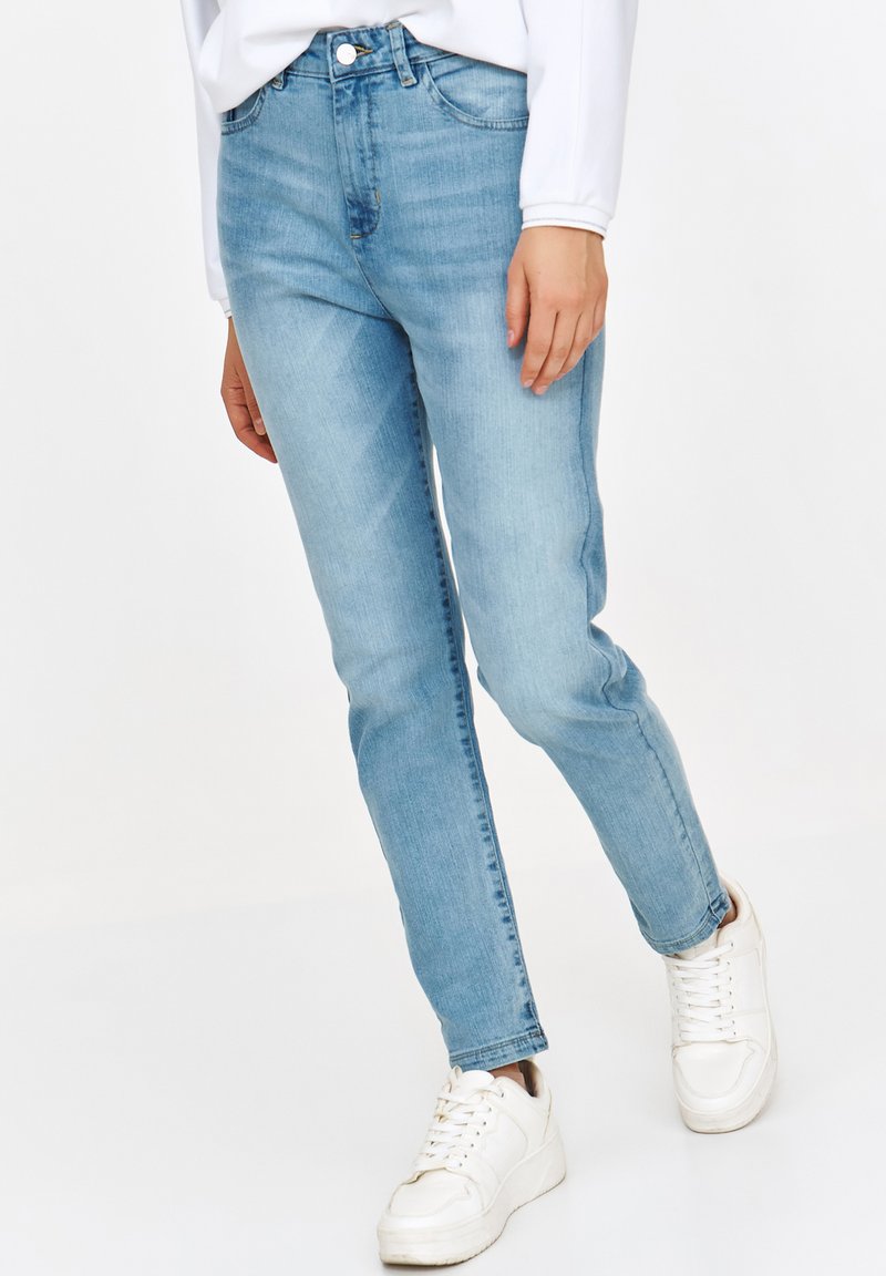 Jasnoniebieskie dżinsy z dopasowanym krojem, z klasycznym pięcioma kieszeniami i zamkiem błyskawicznym. Noszone z białymi trampkami.