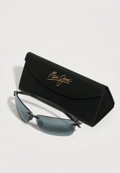 Γυαλιά ηλίου χωρίς πλαίσιο με φακούς με μπλε απόχρωση, τοποθετημένα μπροστά από μια μαύρη πτυσσόμενη θήκη με χρυσό λογότυπο "Maui Jim".